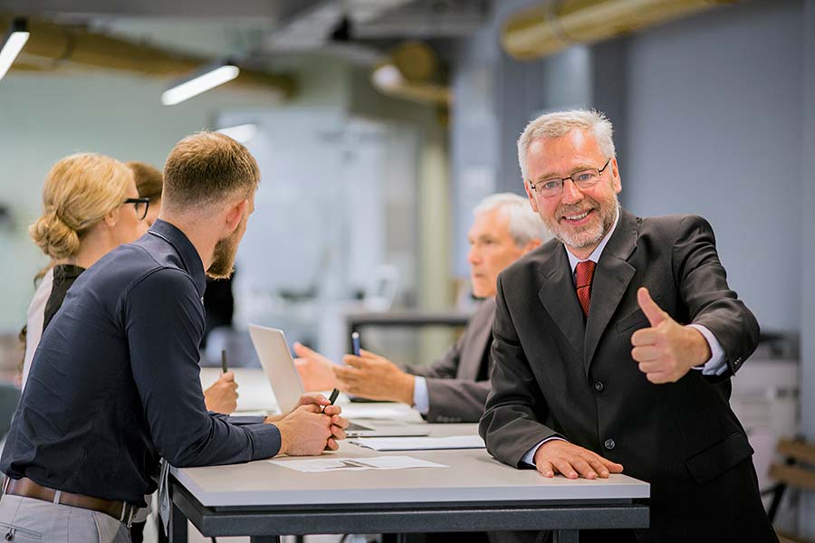Smiling senior businessman showing thumb up sign front business people discussing office