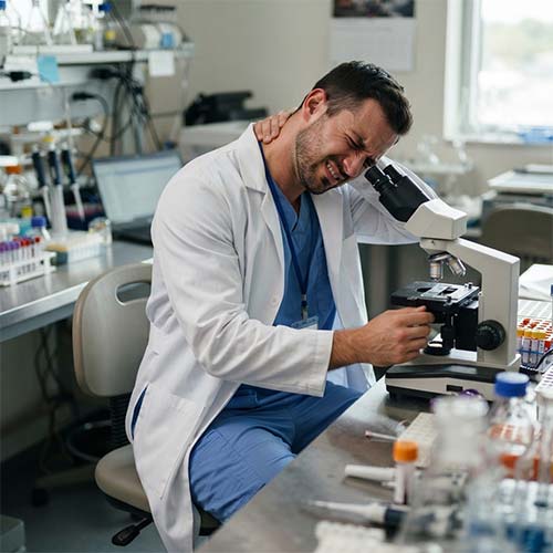 Lab technician holding neck in pain while using microscope, showing repetitive strain injury at workplace.