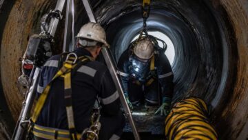 Confined space hazards entry: worker in respirator and harness inside pipe, attendant monitoring at tripod.