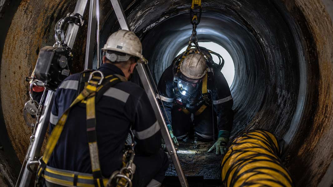 Confined space hazards entry: worker in respirator and harness inside pipe, attendant monitoring at tripod.